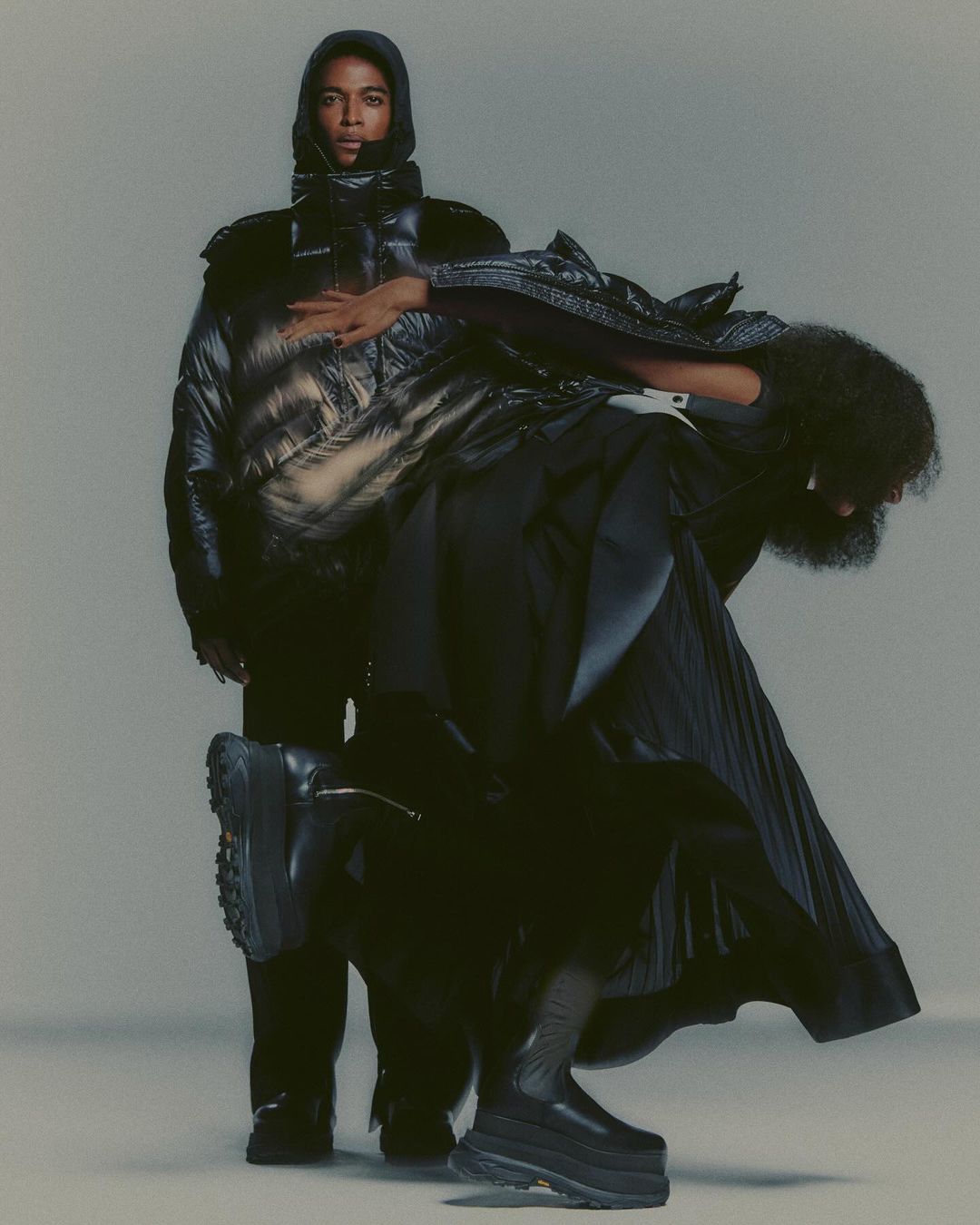 Two fashion models in a studio setting, one standing in a puffer jacket with a hood and another with a long skirt and platform boots.