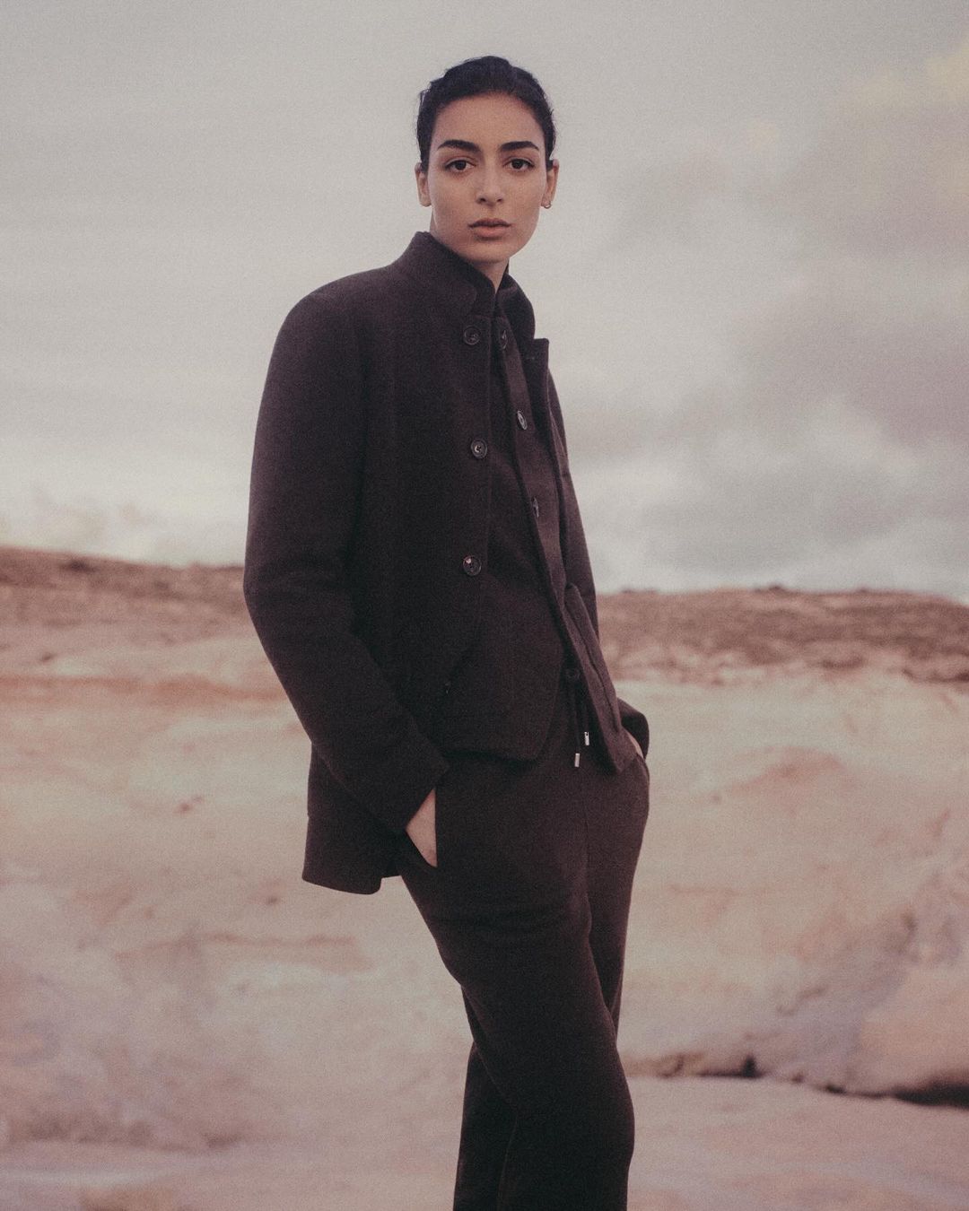 A slender woman poses outdoors wearing a stylish brown jacket and matching trousers, set against a soft, diffused background.