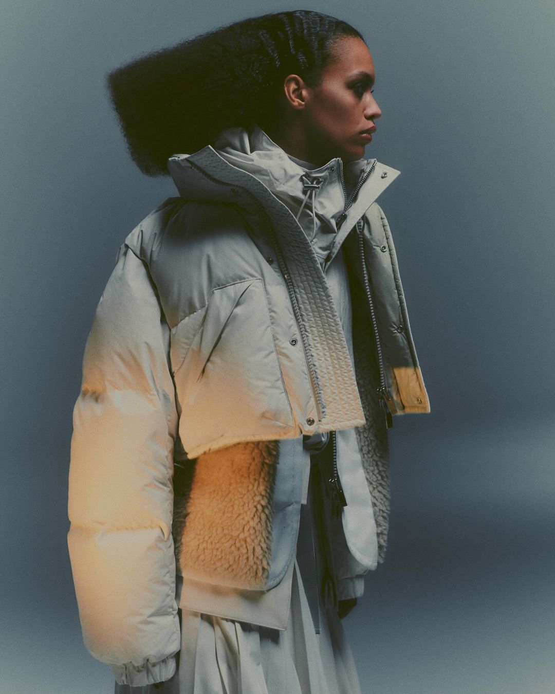 Fashion shot of a woman wearing a gray puffy jacket and a gray skirt, in a studio with gray background and soft lighting.