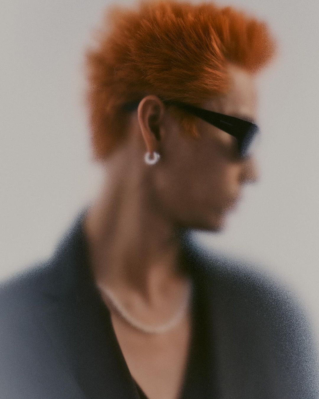 Portrait of a man with bright orange spiky hair wearing sunglasses and a hoop earring against a plain background.