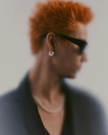 Portrait of a man with bright orange spiky hair wearing sunglasses and a hoop earring against a plain background.