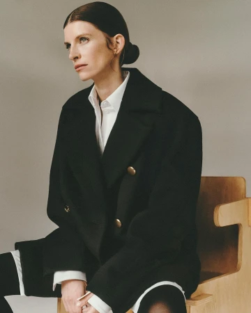 Woman in a black double-breasted coat and white collared shirt seated on a wooden chair against a plain light wall.
