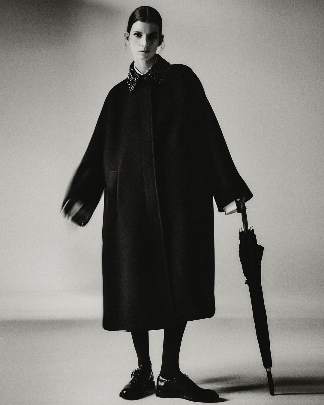 Monochrome studio fashion portrait of an adult woman posing in a dark coat with an umbrella, dark tights, and dark shoes.