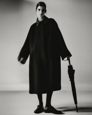 Monochrome studio fashion portrait of an adult woman posing in a dark coat with an umbrella, dark tights, and dark shoes.