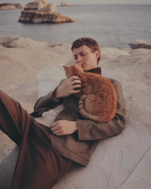 A fair-skinned man wearing brown pants and blazer holds a ginger cat while sitting on rocks near the ocean.