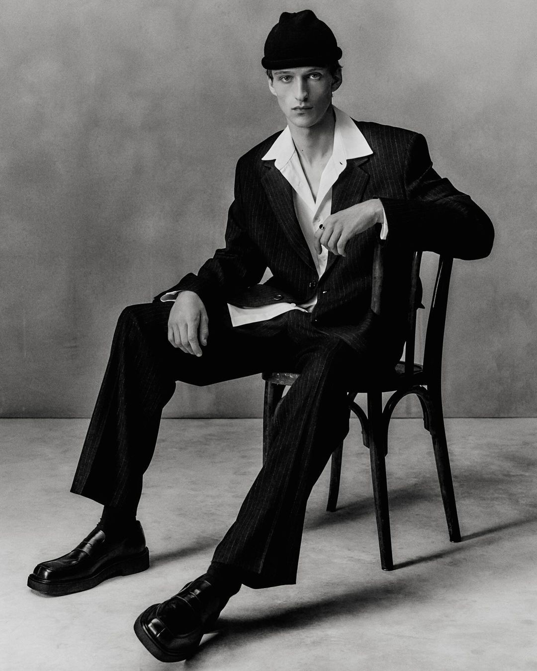 Monochrome portrait of a young man in a dark pinstripe suit and beanie seated on a chair by Darren Gwynn