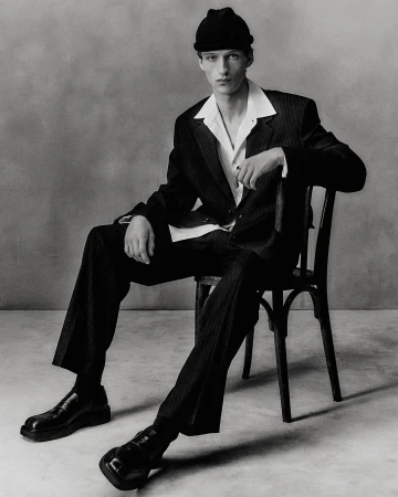 Monochrome portrait of a young man in a dark pinstripe suit and beanie seated on a chair by Darren Gwynn