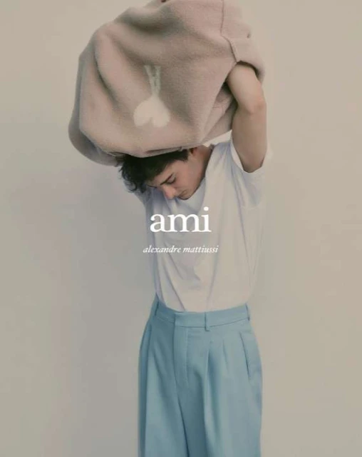 A young man in a white t-shirt and blue pants is taking off a beige jacket with a white heart design in a studio fashion shot.