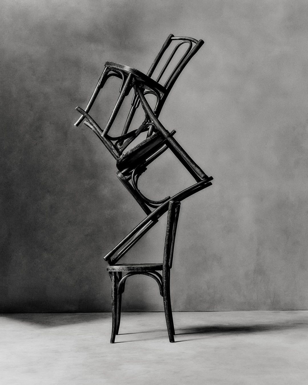 Monochromatic studio shot of three bentwood chairs stacked unevenly, creating an abstract furniture art installation against a gray backdrop.