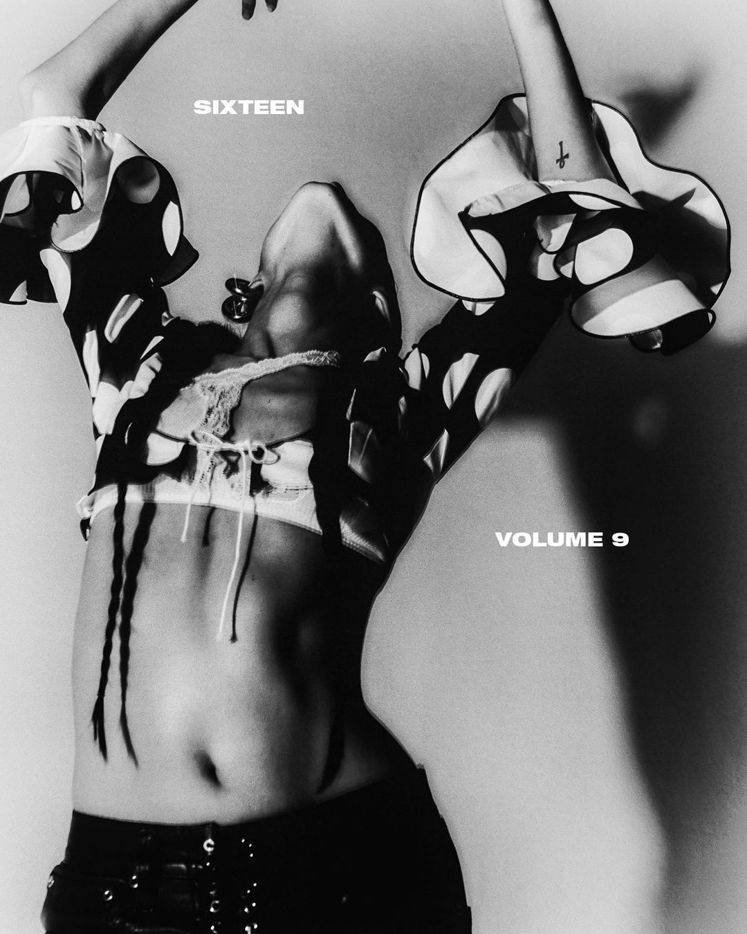 Black and white fashion shot of a person posing with bare torso, black bottoms, jewelry, and a polka dot long sleeve top against a neutral backdrop.
