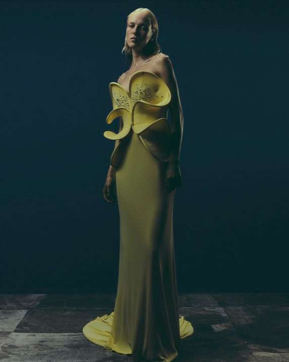 A woman in a floor-length yellow couture gown with abstract floral elements poses against a dark, textured backdrop, fashion-forward.