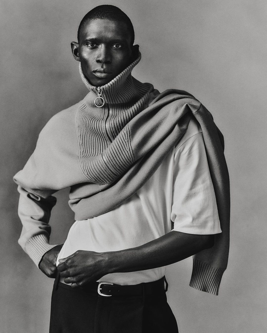 Monochrome portrait of a slender adult man in a white t-shirt and sweater around his shoulders in a studio setting.