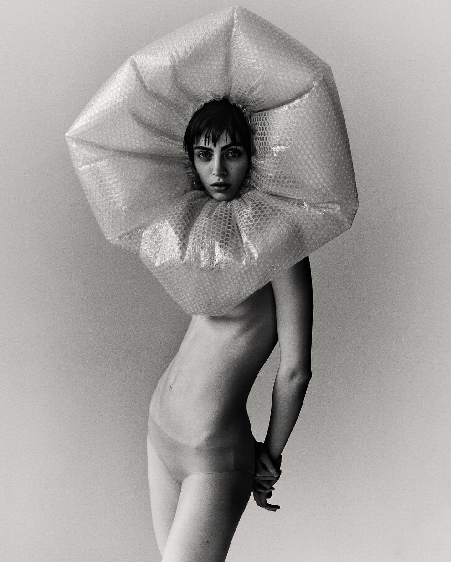 Striking black and white portrait of a woman wearing a bubble wrap collar and headpiece against a plain backdrop, fashion, conceptual.