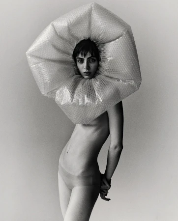 Striking black and white portrait of a woman wearing a bubble wrap collar and headpiece against a plain backdrop, fashion, conceptual.