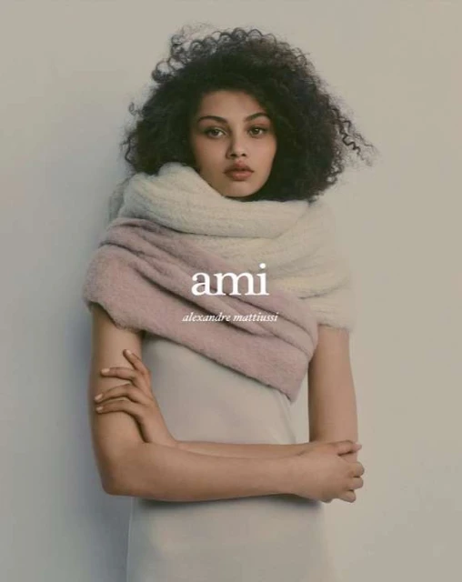 A fashionable adult woman with curly hair, wearing a pale lilac dress and a thick, two-tone scarf, posing indoors against a light background.