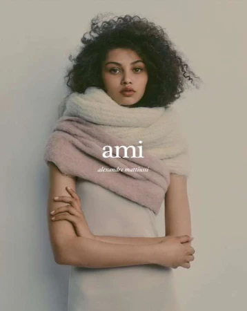 Portrait of a woman with curly hair wearing a thick fluffy scarf and light dress against a neutral grey wall background.