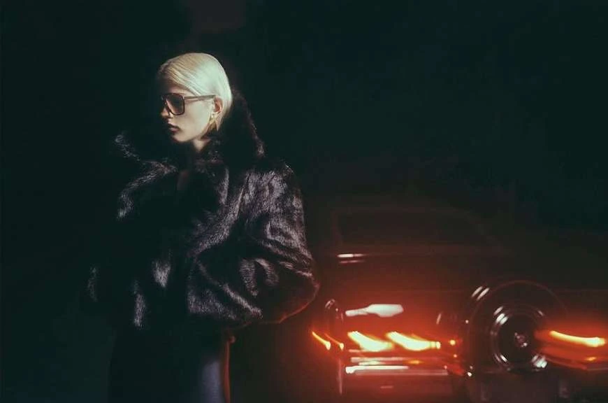 Elegant woman in a fur coat and sunglasses stands next to a classic car in a nighttime setting, lit with a moody, cinematic vibe.