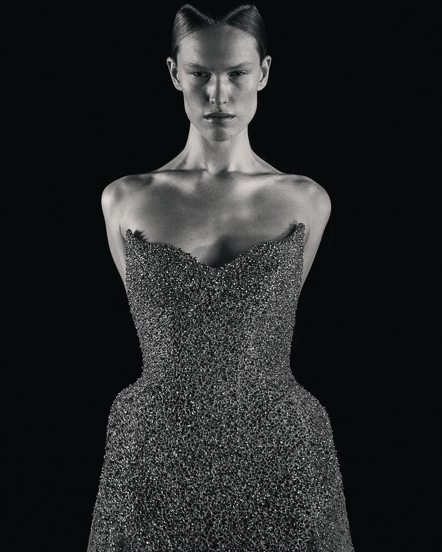 A woman in a beaded strapless gown stands against a stark black backdrop, with her hair styled in an updo and a neutral facial expression.