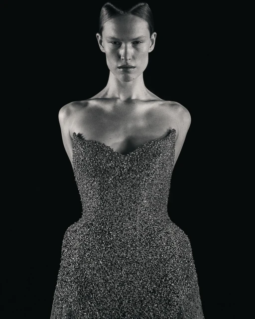 A woman in a beaded strapless gown stands against a stark black backdrop, with her hair styled in an updo and a neutral facial expression.