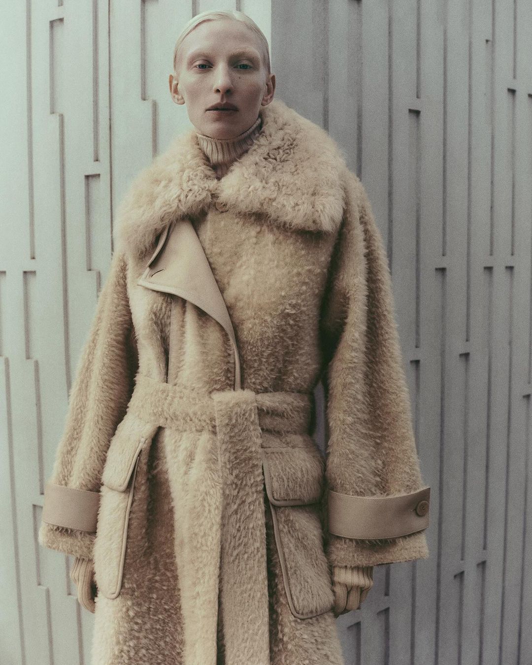 An adult female poses indoors wearing a beige, faux fur coat with matching gloves in front of a textured wall for a fashion editorial.