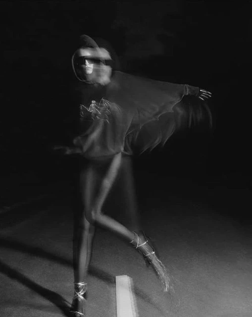 Black and white long exposure photo of a person with legs and arms extended wearing shoes with straps, against dark background.
