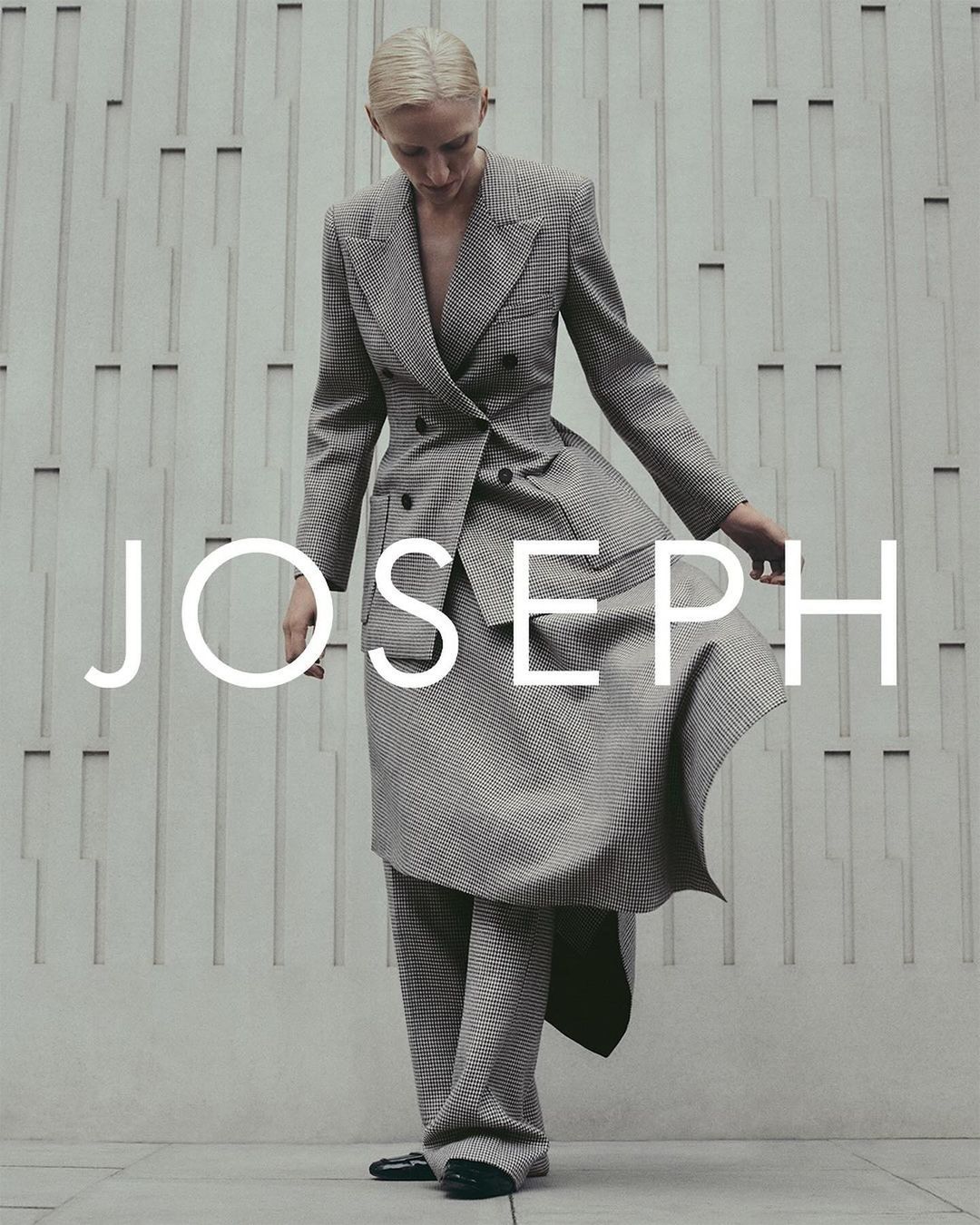 Woman in grey plaid suit, posing against textured wall with JOSEPH logo. Fashion editorial shot, full-length, neutral color palette.