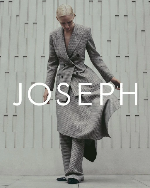 Woman in grey plaid suit, posing against textured wall with JOSEPH logo. Fashion editorial shot, full-length, neutral color palette.