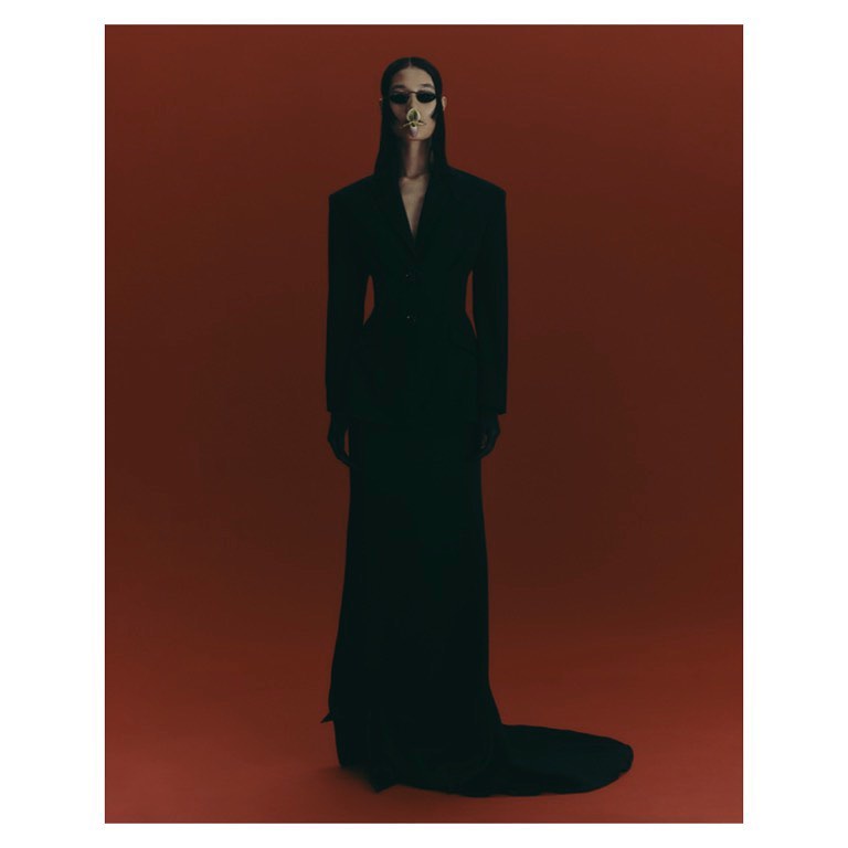 A model in a black dress with a skull-painted face poses in a studio against a dark red backdrop for a fashion editorial photo.