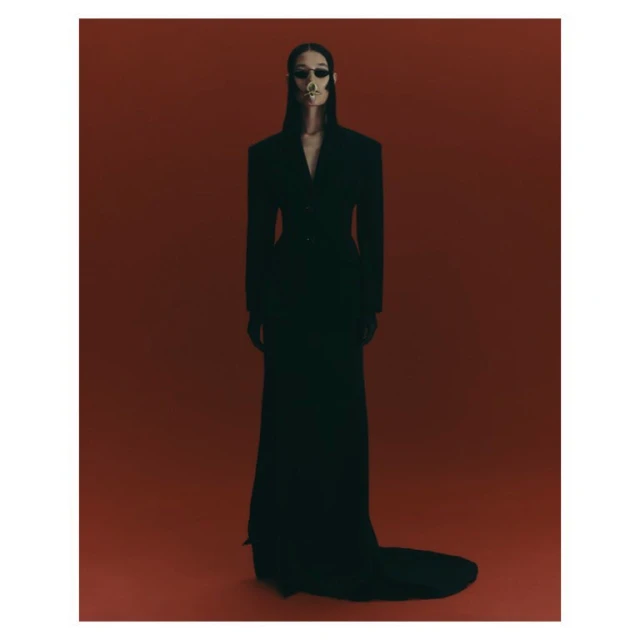 A model in a black dress with a skull-painted face poses in a studio against a dark red backdrop for a fashion editorial photo.