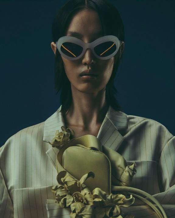 Portrait of a stylish adult woman wearing sunglasses and a striped blazer with a light yellow purse with flower-like decorations.