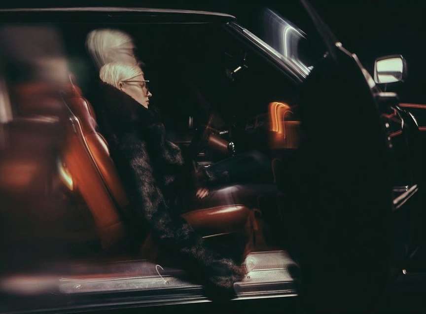 An adult woman with short platinum hair wearing glasses and a fur coat sits in an vintage car with orange leather seats in a dark, moody setting.