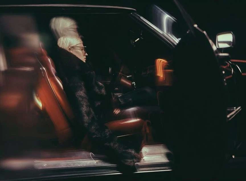 An adult woman with short platinum hair wearing glasses and a fur coat sits in an vintage car with orange leather seats in a dark, moody setting.