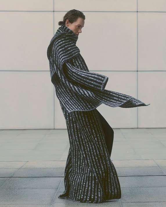 Fashion shot of a model in a black and silver striped dress with a high neck, posing in an indoor setting with natural light.
