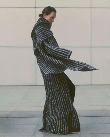 Model in dark striped ribbed knit coat with matching scarf standing against large tiled architectural wall