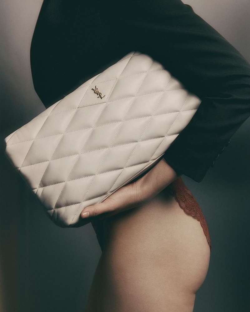 Close-up studio shot featuring a cream-colored, quilted Saint Laurent clutch purse held by a fair-skinned woman wearing black and red clothing.