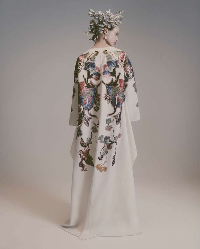 Model wearing an off-white dress with floral back detailing and a floral headpiece in a light grey studio setting looking off camera.