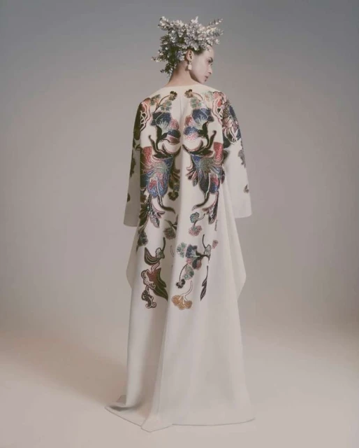 Model wearing an off-white dress with floral back detailing and a floral headpiece in a light grey studio setting looking off camera.