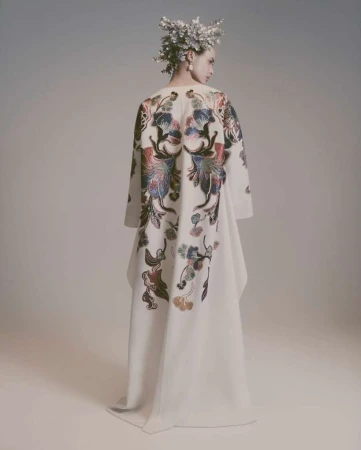 Model wearing an off-white dress with floral back detailing and a floral headpiece in a light grey studio setting looking off camera.