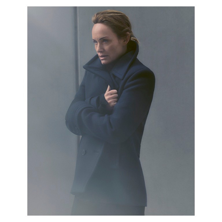 Woman in a navy coat with arms crossed stands against a gray background, creating a minimalist fashion portrait with natural lighting.