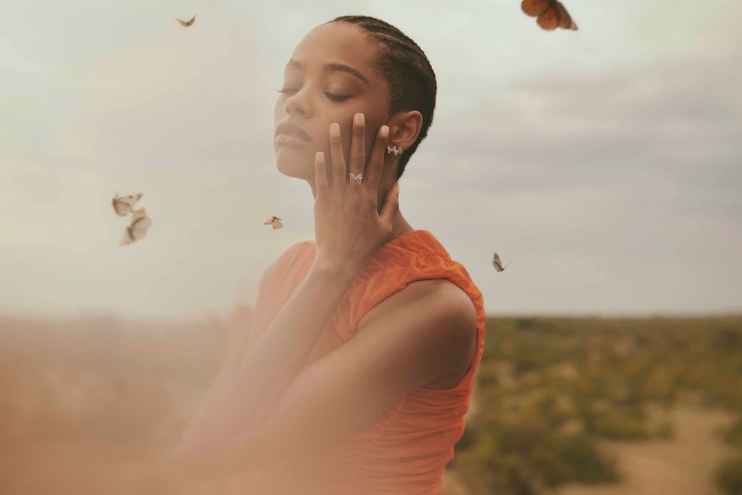 Woman with short hair in an orange dress posing in a field with flying butterflies, wearing butterfly jewelry with a soft, dreamy aesthetic.