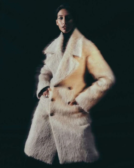 Elegant woman in a cream-colored, furry coat poses in a studio with dark background and soft lighting for a fashion editorial shoot.