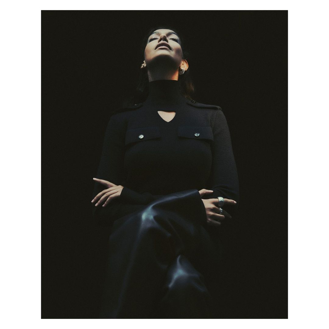 A woman in a dark turtleneck sweater, with her head tilted back, stands in a dimly lit space, creating a moody, sophisticated portrait.