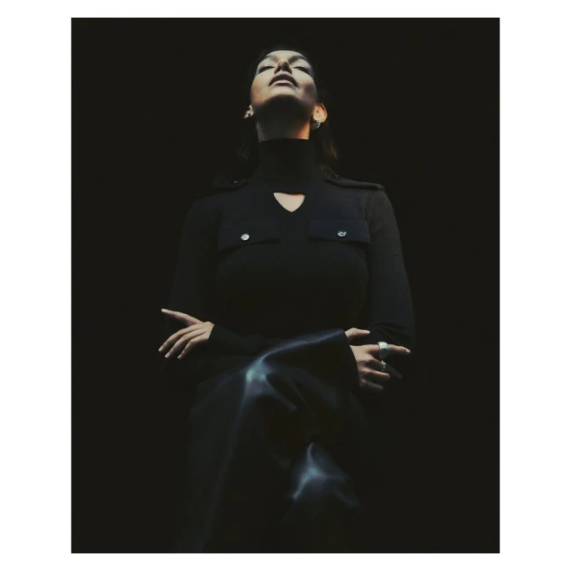 A woman in a dark turtleneck sweater, with her head tilted back, stands in a dimly lit space, creating a moody, sophisticated portrait.