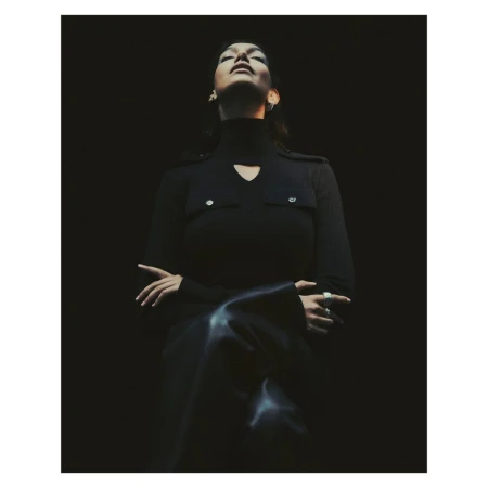 A woman in a dark turtleneck sweater, with her head tilted back, stands in a dimly lit space, creating a moody, sophisticated portrait.