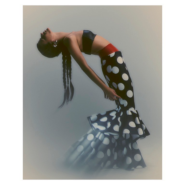 Woman in a black and white polka dot dress arches her back in a studio setting with diffused lighting, posing in a fashion forward style.
