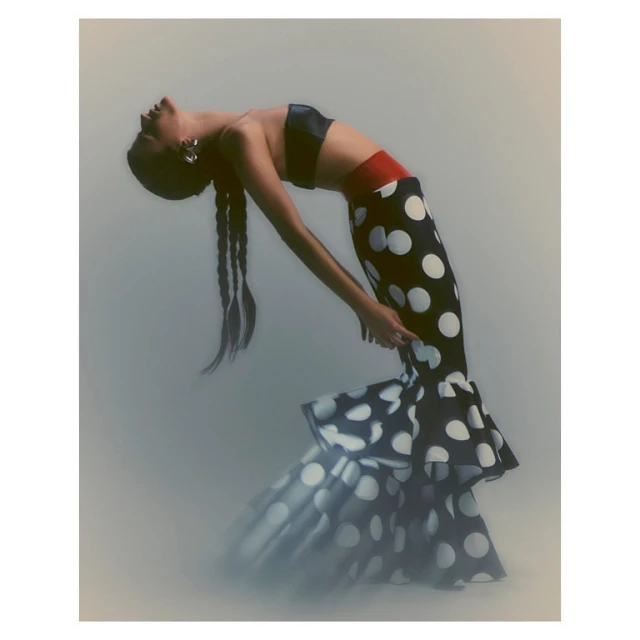 Woman in a black and white polka dot dress arches her back in a studio setting with diffused lighting, posing in a fashion forward style.