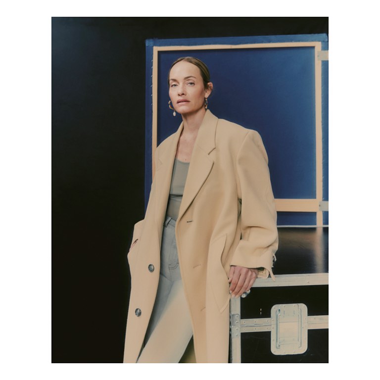 A woman in a beige coat poses in front of a blue backdrop with a metal frame. She is wearing a neutral top and light jeans.