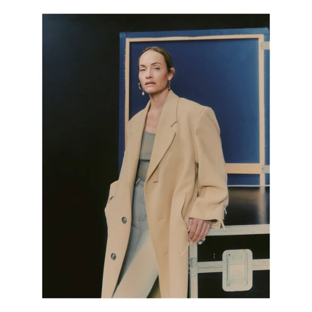 A woman in a beige coat poses in front of a blue backdrop with a metal frame. She is wearing a neutral top and light jeans.