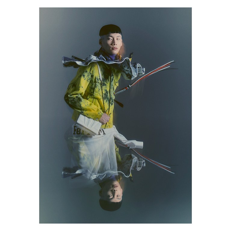Fashionable Asian male model in stylized yellow jacket holds white book, with reflective surface creating a striking mirrored effect.