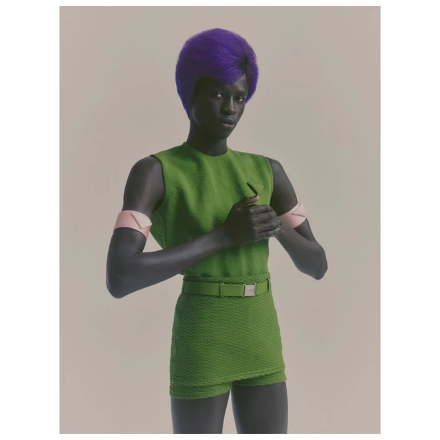 A striking portrait of an adult woman with a bright purple afro, wearing a green textured dress with matching belt, set against a neutral backdrop.